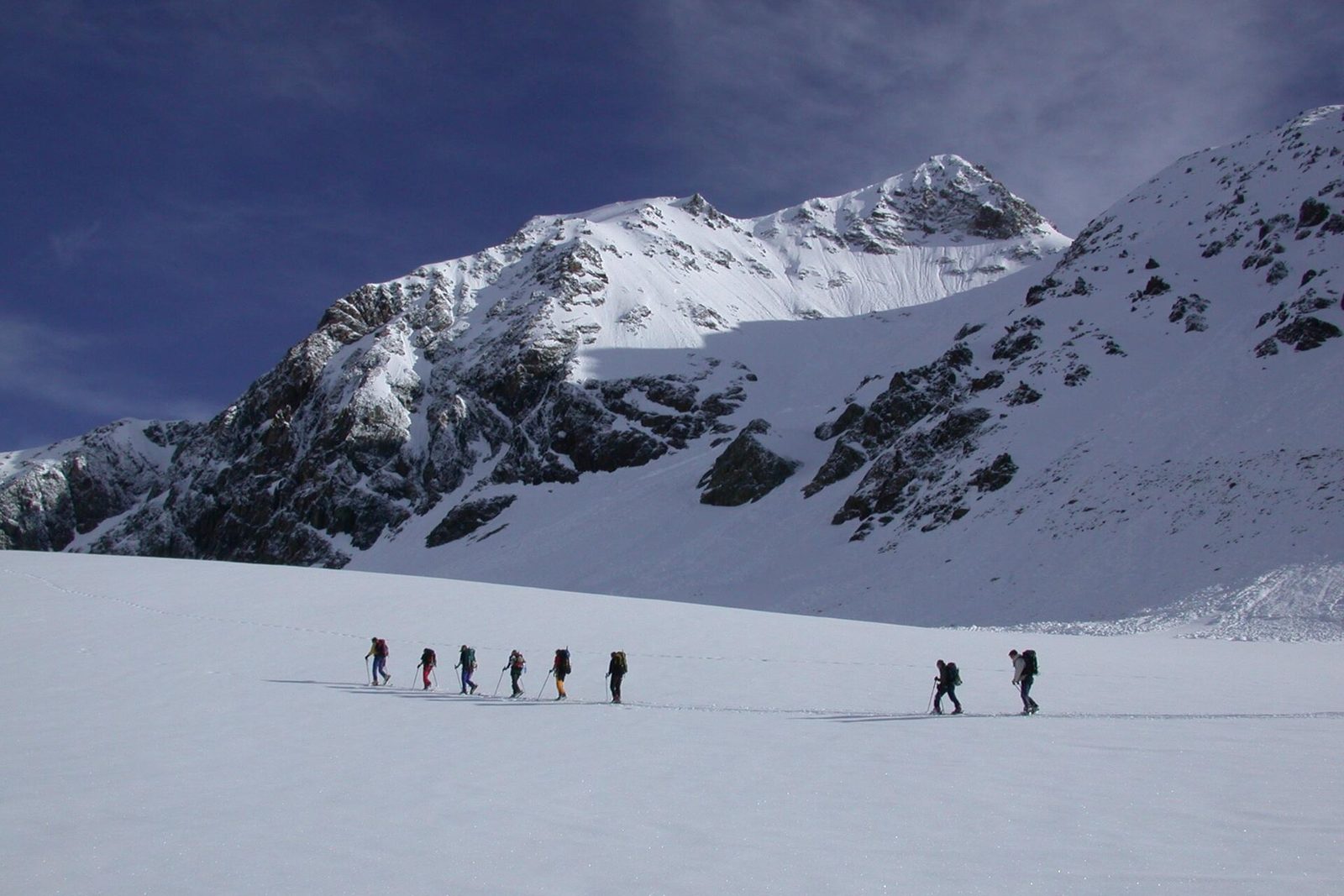 A line of ski tourers crossing a snowfield below a rock face